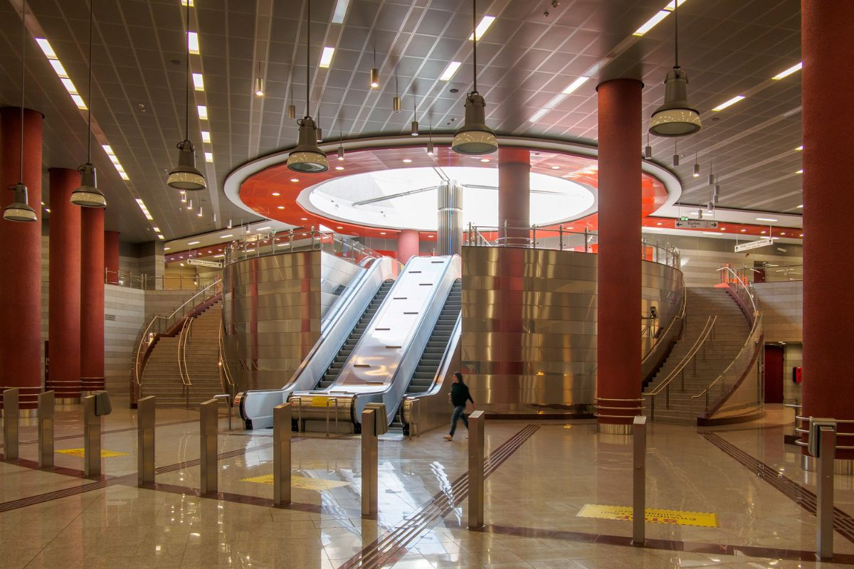 A modern subway station interior with escalators, stairs, red columns, and hanging lights.